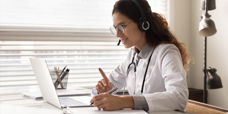 A female doctor takes notes as she consults a patient via telehealth