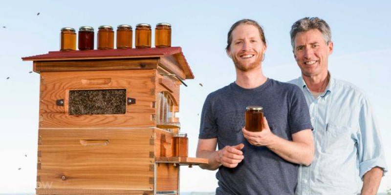 Two men stand next to a bee hive holding a jar of honey