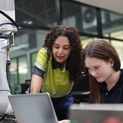 Two women, working in a manufacturing setting, look at a laptop screen.
