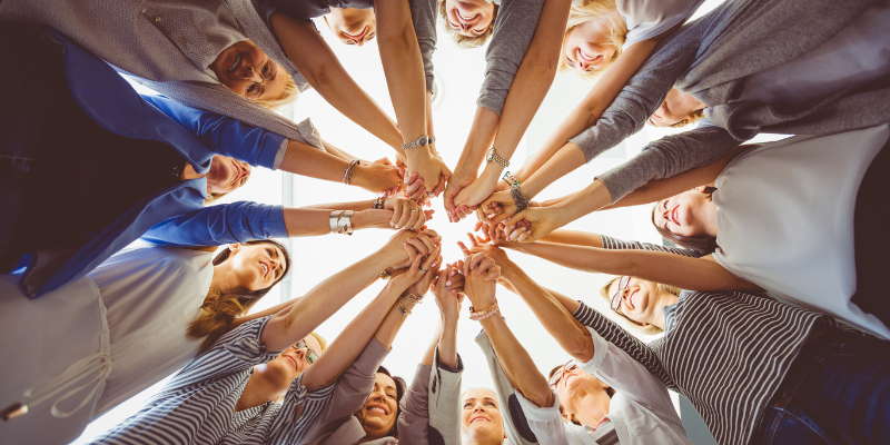 an image of a group of women standing in a circle, their hands outreached in the shape of a star. The image is photographed from below. 