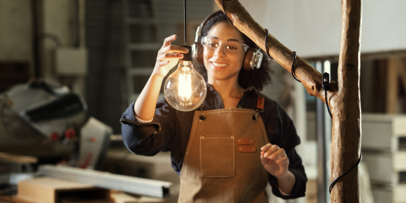 A person in a workshop holding up a glowing light bulb beside a wooden structure.