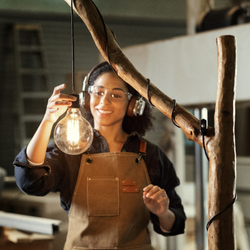 A person in a workshop holding up a glowing light bulb beside a wooden structure.