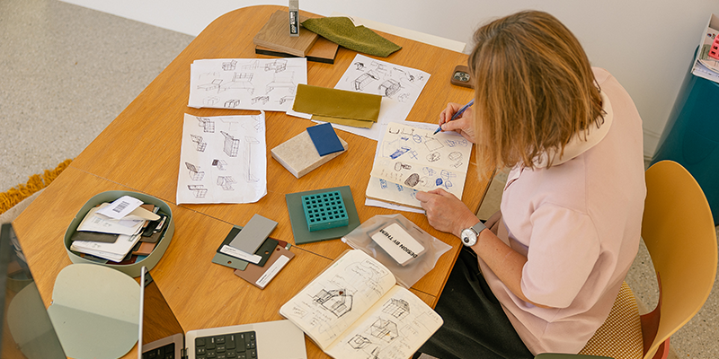 Overhead view of a person sketching product design concepts in a notebook at a desk covered with prototypes, material samples, and design drawings.