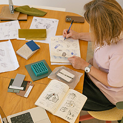 Overhead view of a person sketching product design concepts in a notebook at a desk covered with prototypes, material samples, and design drawings.