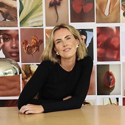 Person with short light hair wearing a black top, seated in front of a grid wall filled with colourful images.