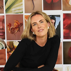 Person with short light hair wearing a black top, seated in front of a grid wall filled with colourful images.