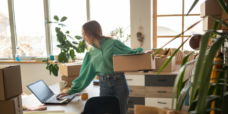 Caucasian entrepreneur holds box in left hand while replying to email in business studio