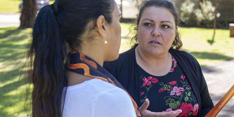 The side profile of an indigenous woman speaking to another woman who is holding documents and folders in her hand. They are standing outside. 