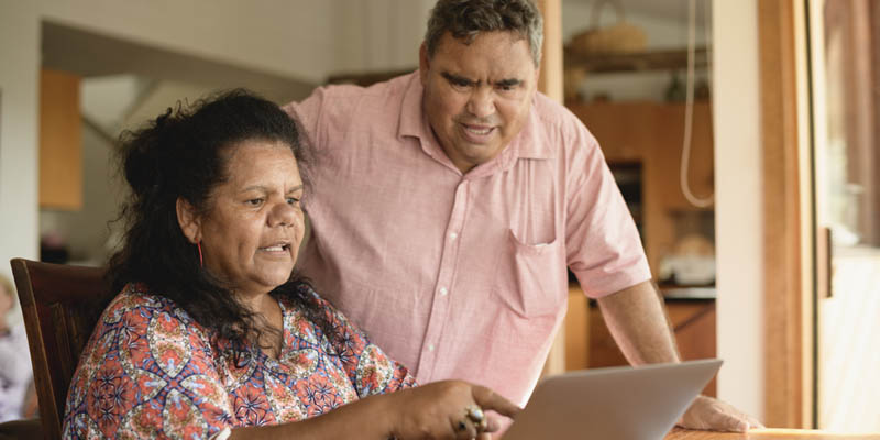 Australian Indigenous woman sitting in front of a laptop. She is explaining something to an indigenous man who is standing next to her. 