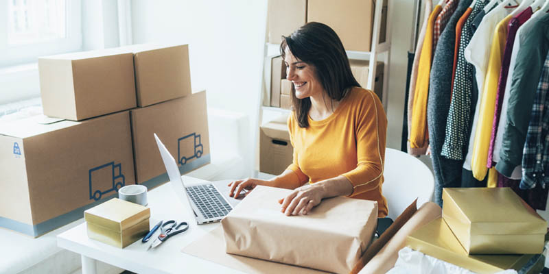 A woman sitting at a desk preparing packages to send to clients. She is entering information on her laptop. There are packing boxes and a rack of clothes in the background. 