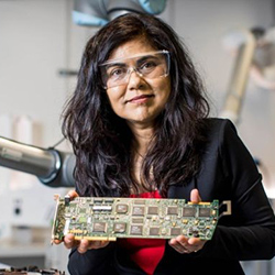 Professor Veena Sahajwalla AO wearing safety glasses and holding an electronic circuit board in a lab.