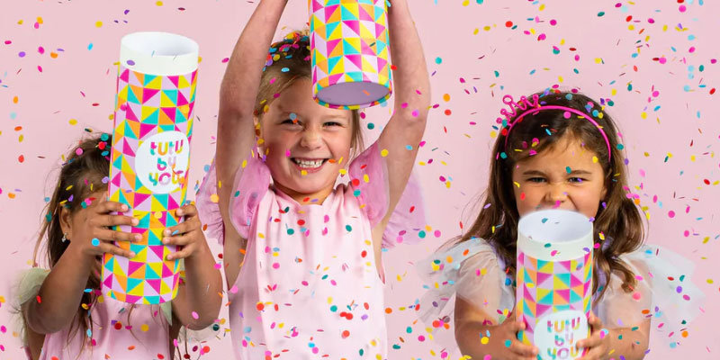 Three little girls dressed in party clothes, hold colourful branded 'Tutu by You' cylinders. They're surrounded by mid-air confetti with a pink background